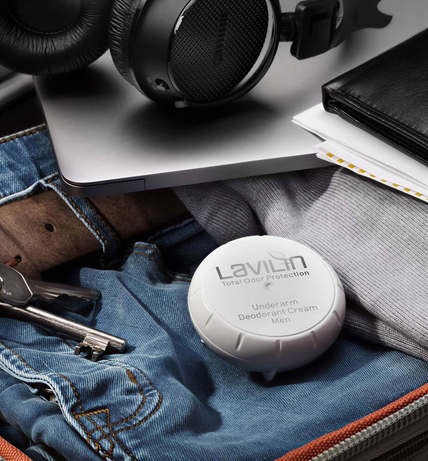 Underarm deodorant cream for men in a white container, placed on denim jeans with headphones and a laptop.